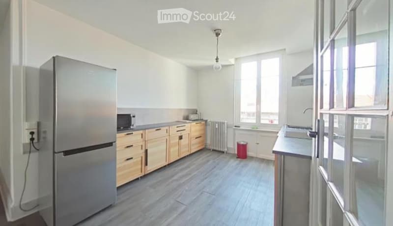 Modern kitchen with light wooden cabinets, stainless steel refrigerator, microwave, sink, and radiator. The space is well-lit with large windows and a grey tiled floor.