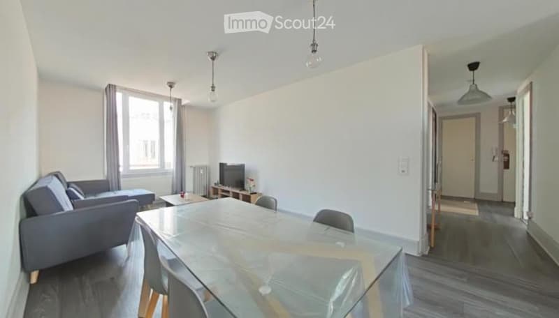 Modern, well-lit living and dining area with a grey sofa, coffee table, TV stand, and a glass dining table with chairs.