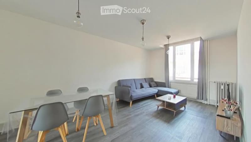 Modern living room with a grey sofa, coffee table, glass dining table, and chairs. Large windows with curtains and pendant lights.