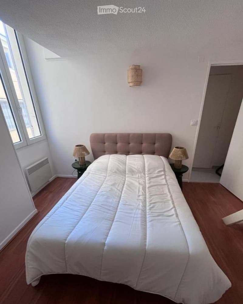 A cozy bedroom with a neatly made bed, two nightstands with lamps, and a wall-mounted light fixture. The room features wooden flooring and large windows letting in natural light.