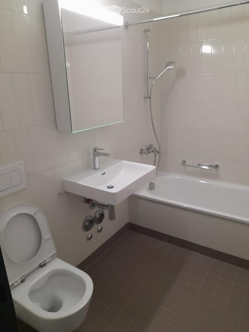 Modern bathroom with a toilet, sink, bathtub, mirror, and showerhead. The walls are tiled, and the floor is covered with brown tiles.