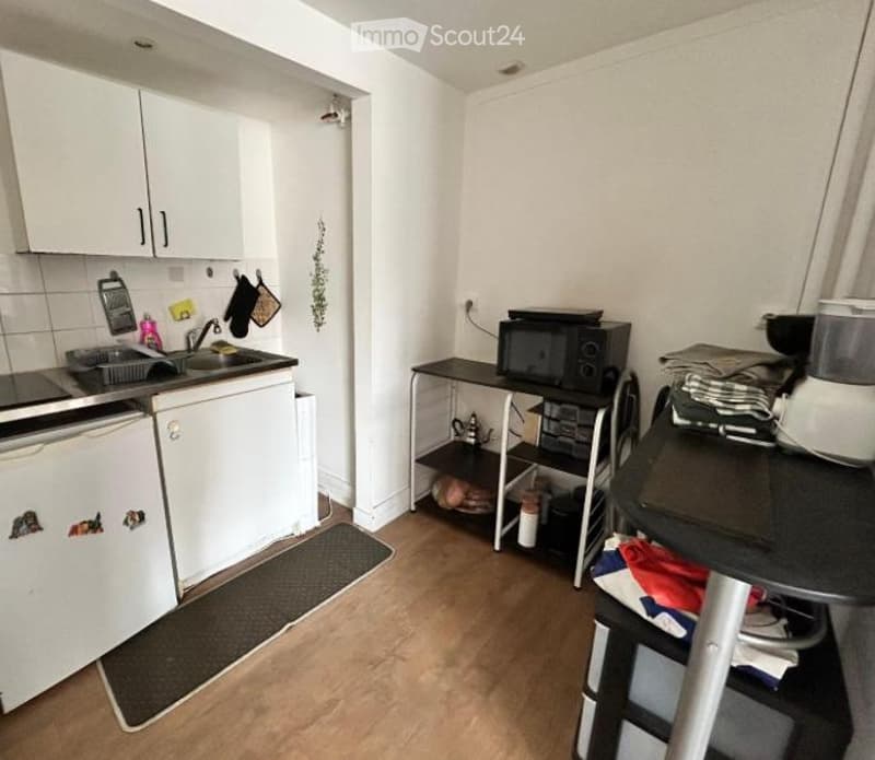 Compact kitchen with white cabinets, a sink, a dishwasher, a microwave, and a small table with a chair. The floor is wooden, and there are various kitchen items and utensils visible.