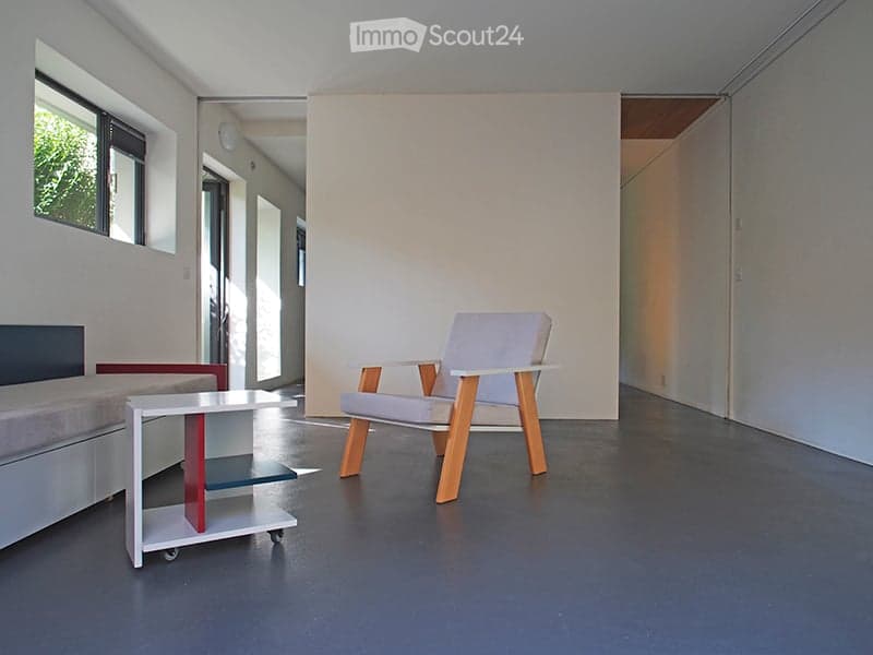 A modern, minimalist room with a grey sofa, a white wooden chair, and a small white table with red accents. The room features large windows and a clean, contemporary design.