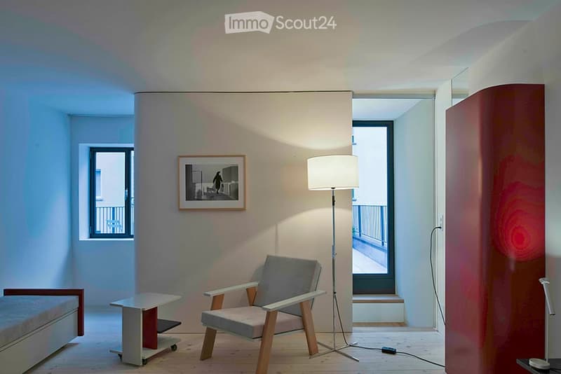 A modern, minimalist room with a grey upholstered chair, a small white table, a floor lamp, and a framed picture on the wall. The room features light wooden flooring and large windows with a view of a balcony.