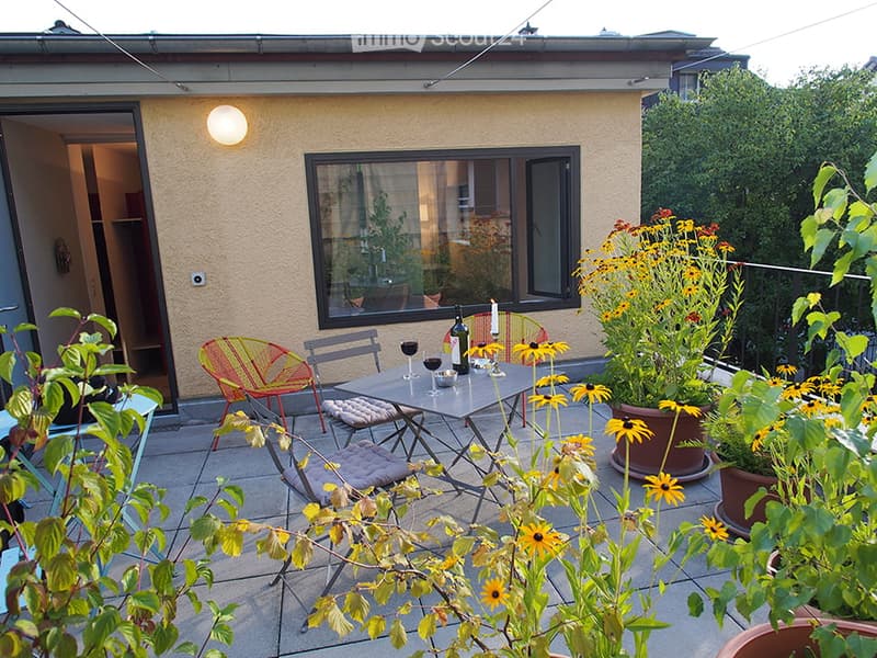 Balcony with a small table, chairs, and potted flowers, adjacent to a building with a large window and a lit wall light.
