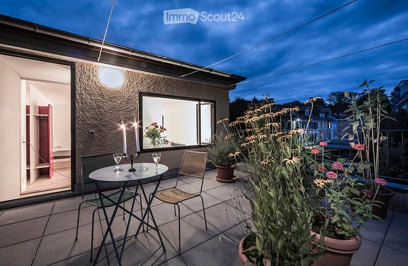 Balcony with a round table, two chairs, candle holder, and potted plants, overlooking a cityscape at dusk.