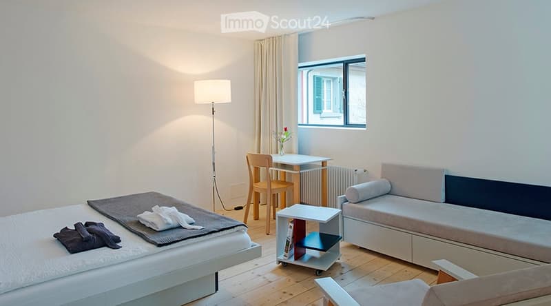 A modern, well-lit room with a bed, sofa, small table, and chair. The room features wooden flooring, white walls, and a large window with curtains. A floor lamp and a small side table with books are also present.
