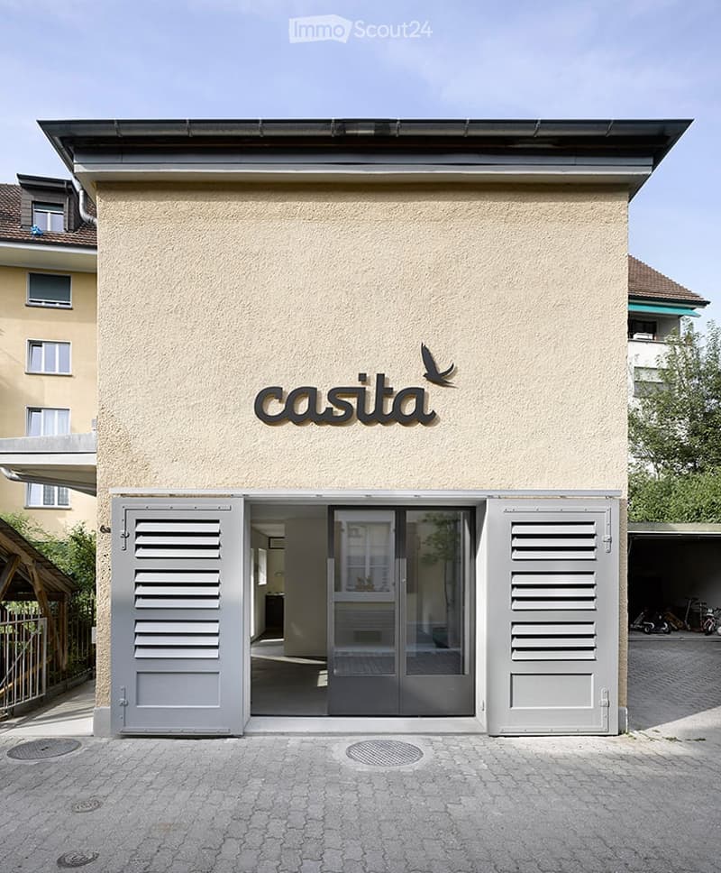 Modern building entrance with a sign reading "casita" above the door, flanked by grey shutters, with a paved area and surrounding greenery.
