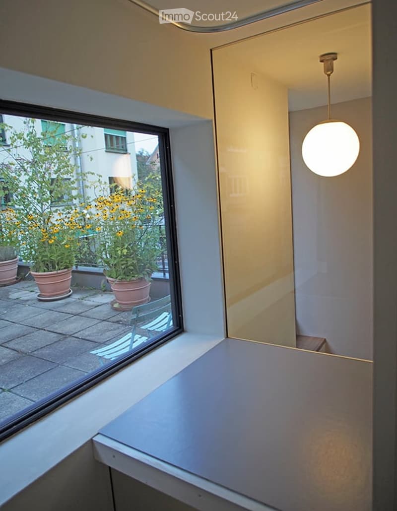 A small, modern room with a large window overlooking a balcony area with potted plants. The room features a simple white table and a single chair, with a modern pendant light hanging from the ceiling.