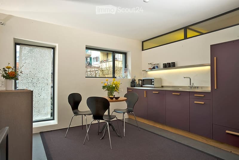 Modern kitchen and dining area with dark purple cabinets, a small dining table with four chairs, and a microwave on the counter. There are two windows with frosted glass and a view of the street.