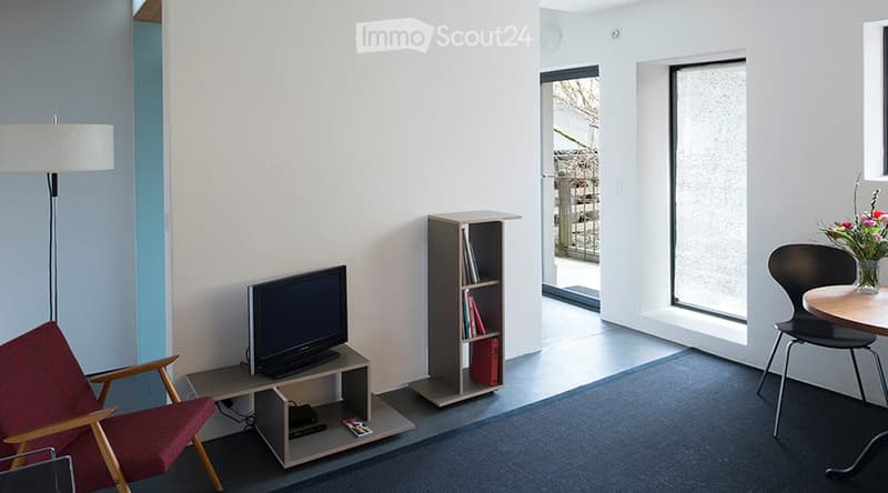 A modern, minimalist room with a red armchair, a small table with a TV, a bookshelf, and a floor lamp. The room features large windows and a view of an outdoor area.