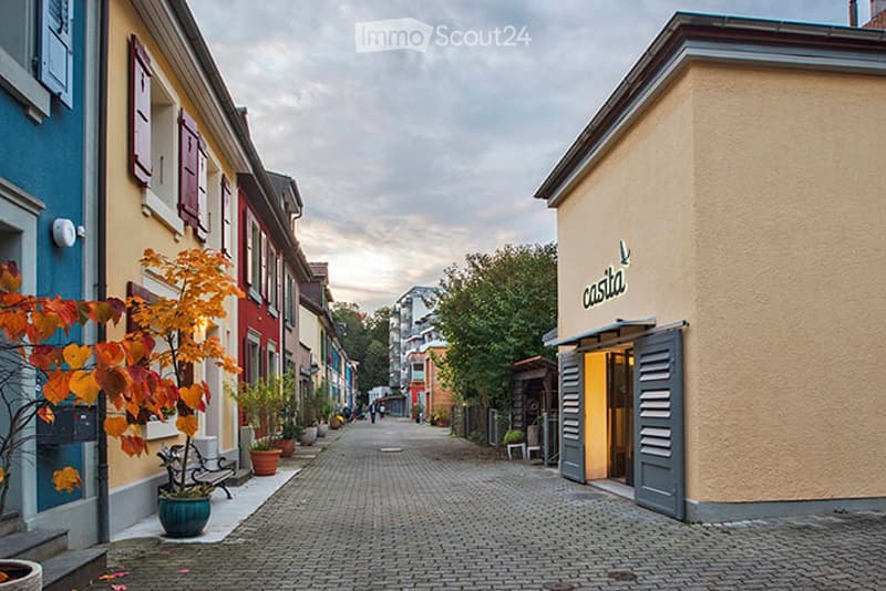 A charming cobblestone street lined with colorful buildings, featuring potted plants and trees, with a shop named "castia" visible on the right.