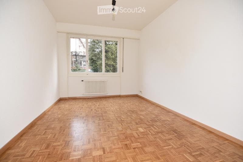 Empty room with wooden parquet flooring, white walls, large window with view, radiator, and ceiling light fixture.