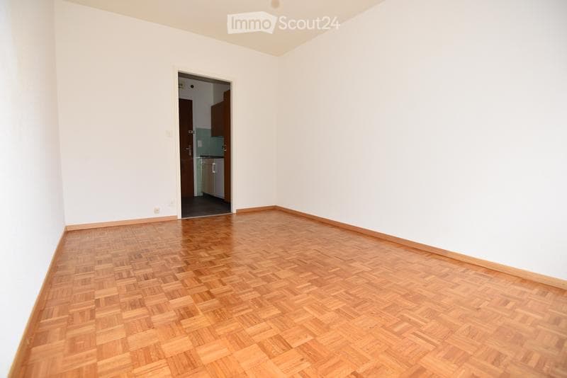 Empty room with wooden parquet flooring and white walls, featuring a doorway leading to another room.