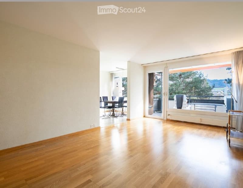 Bright, open living space with wooden flooring, large sliding glass doors leading to a balcony, and a small dining area with a table and chairs.