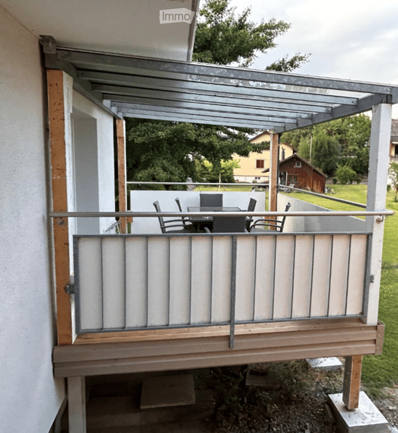 Balcony with a metal roof, wooden decking, and a small dining set including a table and chairs. The view shows a green lawn and trees.