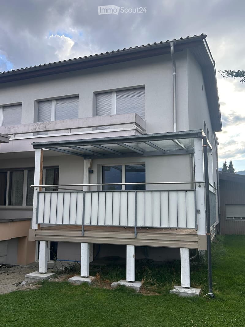 Two-story house with white exterior, multiple windows with blinds, and a raised ground floor with a covered balcony area.