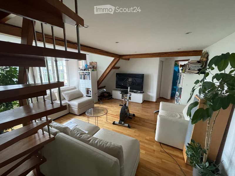 Living room with wooden floors, exposed wooden beams, large windows, and a staircase leading to an upper level. Features a grey sectional sofa, a white armchair, a glass coffee table, an exercise bike, a TV, a bookshelf, and a large potted plant.