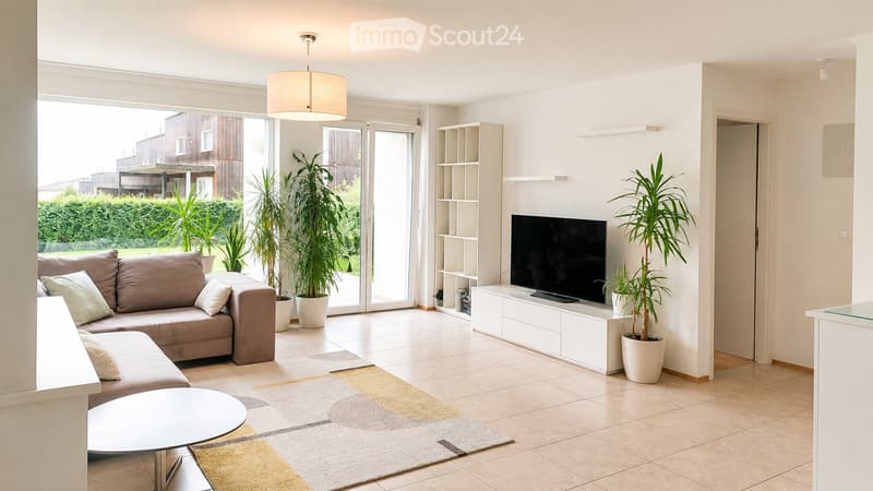 Bright living room with large sliding glass doors leading to a patio, a beige sectional sofa, a white coffee table, a flat-screen TV on a white entertainment unit, and several potted plants.