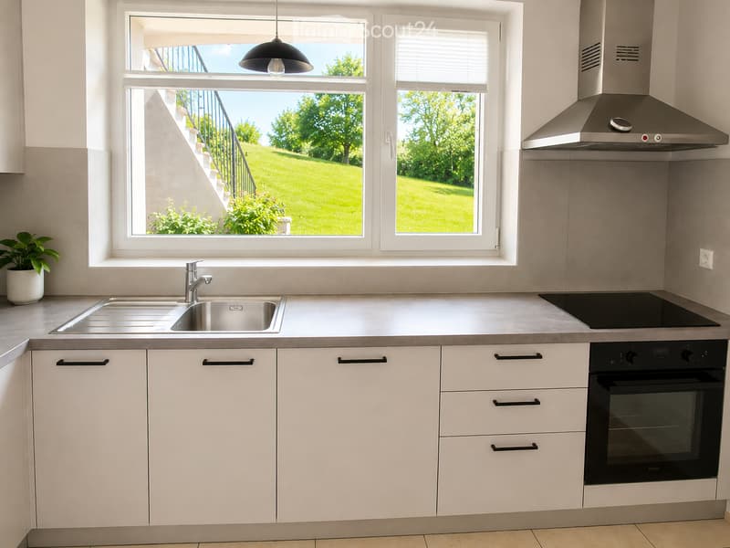 Modern kitchen with white cabinets, stainless steel sink, oven, induction cooktop, and range hood. Large windows offer a view of a green lawn and garden.