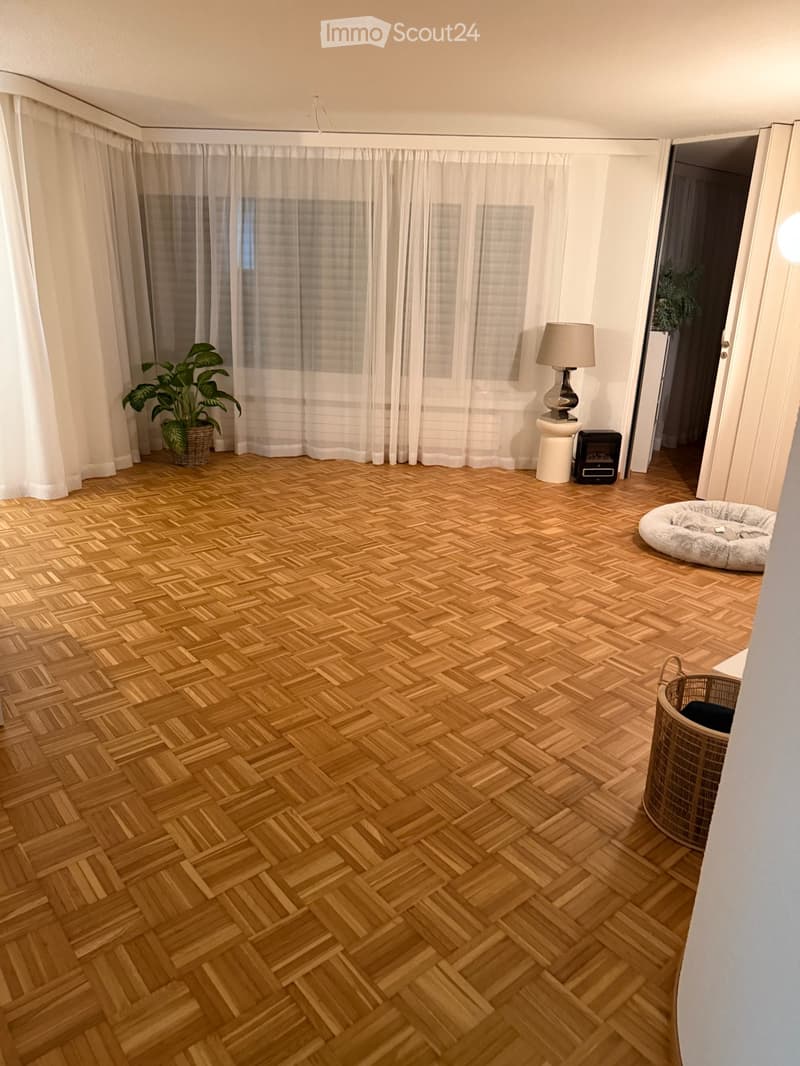 Empty room with wooden parquet flooring, white curtains, a white lamp on a small pedestal, and some potted plants.