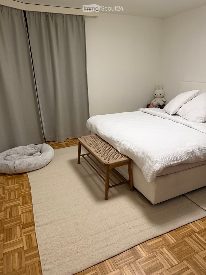 A large bed, white sheets, white mattress, pillows, a bench, rug, and a curtain in front of the bed.