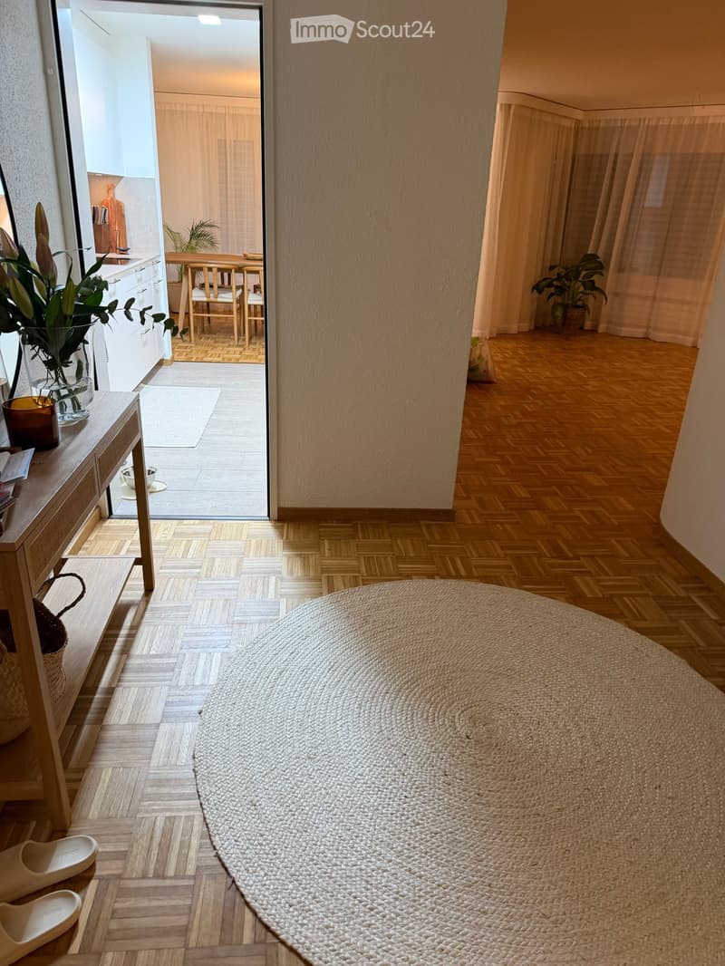entryway with wooden floor, round woven rug, wall-mounted mirror, table with vase, shoe rack, doorway leading to kitchen