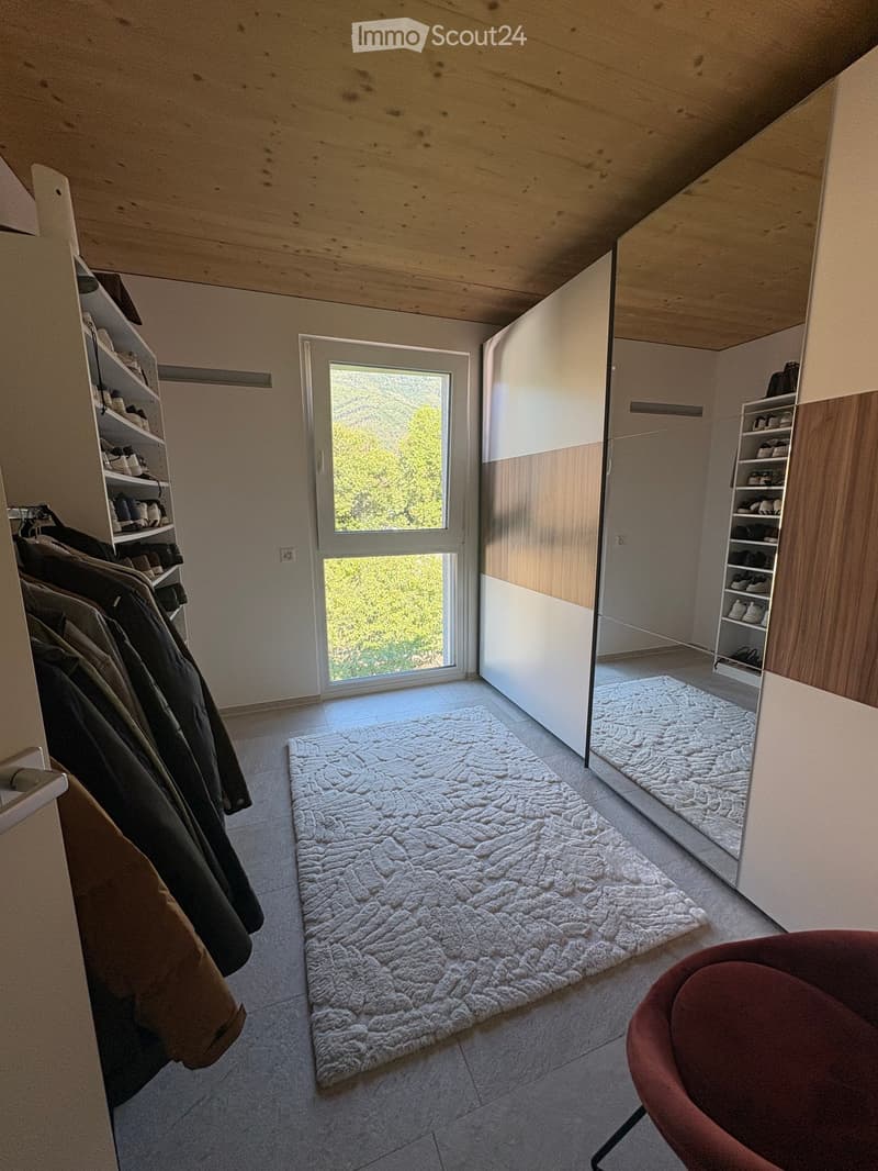 Modern walk-in closet with wooden shelves, clothing racks, a large white shag rug, and a red chair. Features a large window with a scenic view.