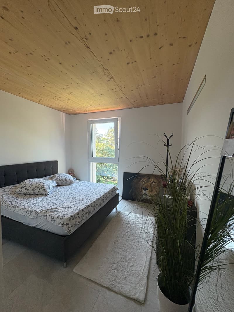 Bright bedroom with a wooden slanted ceiling, a large window showing greenery, a bed with patterned bedding, a white rug, and a potted plant near the window.