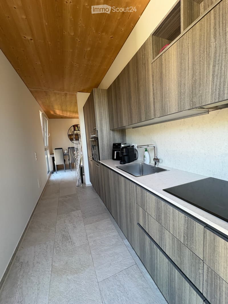Modern hallway leading to a kitchen area with sleek wooden cabinets, a sink, and a coffee machine on the counter. The hallway has a sloped ceiling and light-colored tiles.