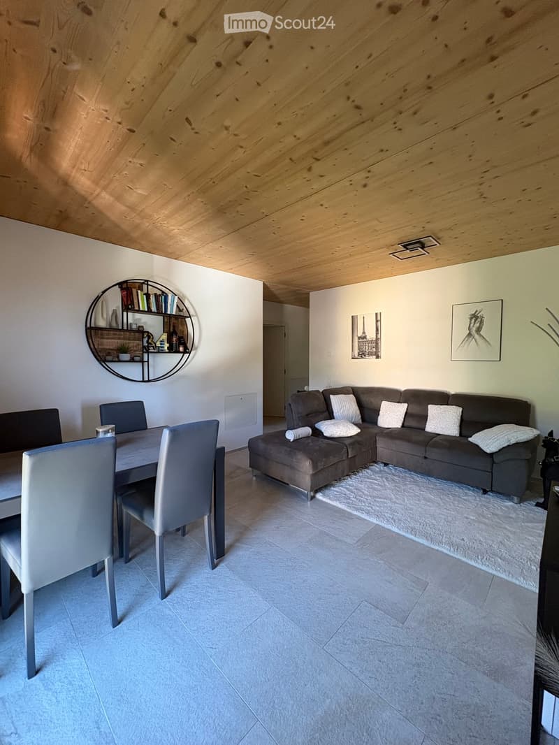 Modern living and dining area with a grey sectional sofa, white throw pillows, a dining table with four chairs, a round wall-mounted shelf, and artwork on the walls.