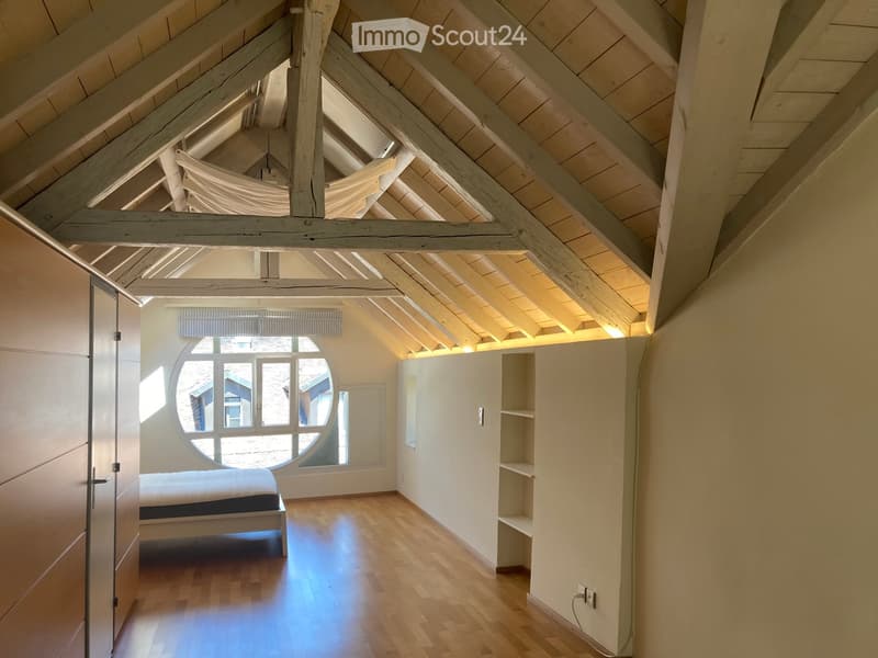 Bright attic bedroom with wooden floors, exposed wooden beams, large round window, built-in shelves, and a simple bed.