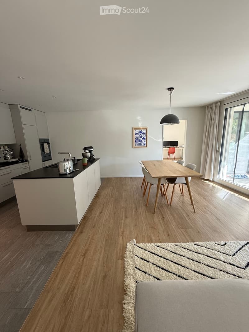 Open-plan living and dining area with light wood flooring, white kitchen cabinets, a central island, a dining table with chairs, a sofa, and a large window with curtains.