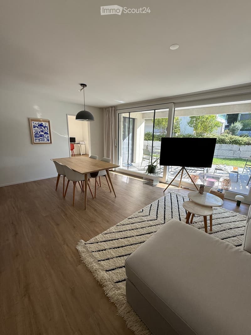Modern living room with wooden flooring, large windows, a sofa, coffee table, and a dining area with a table and chairs.