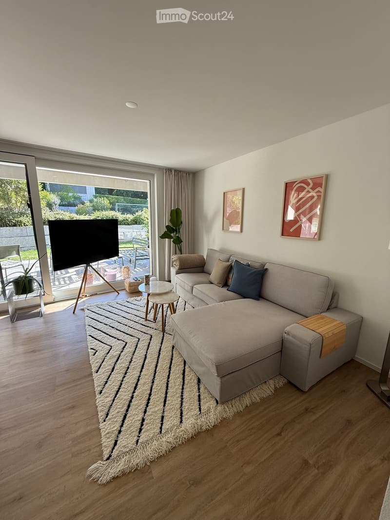 Modern living room with a sectional sofa, coffee table, small stool, television on a stand, and a large area rug. The room features large windows with curtains and a view of a garden.