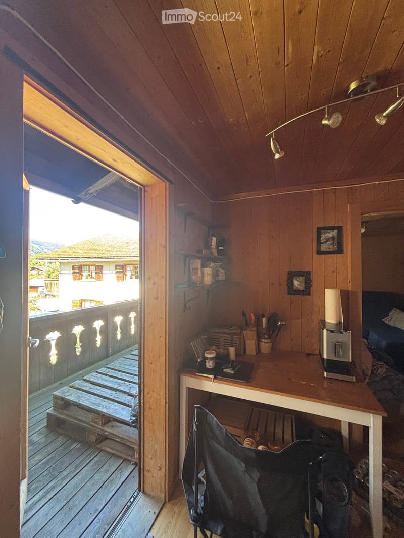 Cozy attic room with wooden walls and ceiling, featuring a small desk, chair, and shelves. An open door leads to a wooden deck with a scenic view.