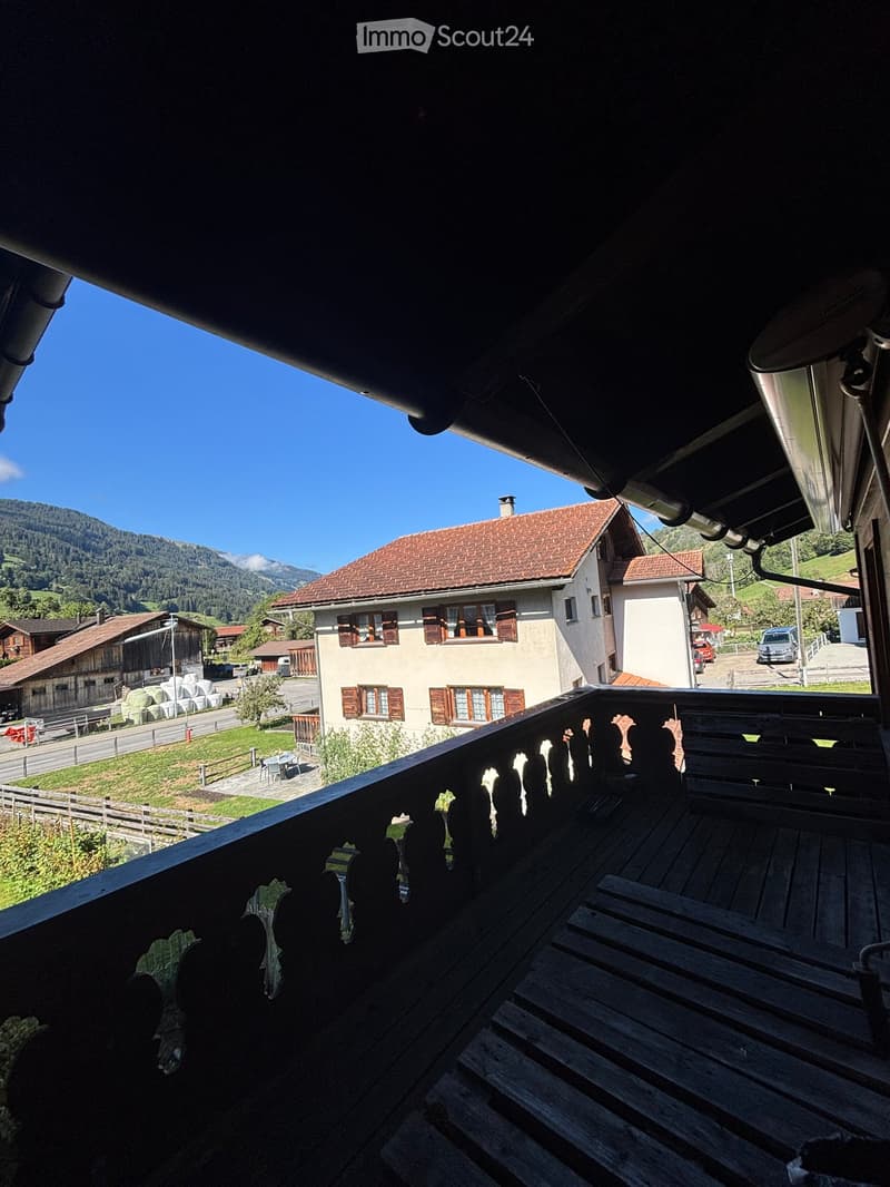 Wooden balcony with railing overlooking a rural village and mountains, with a few parked cars and a small table visible in the distance.