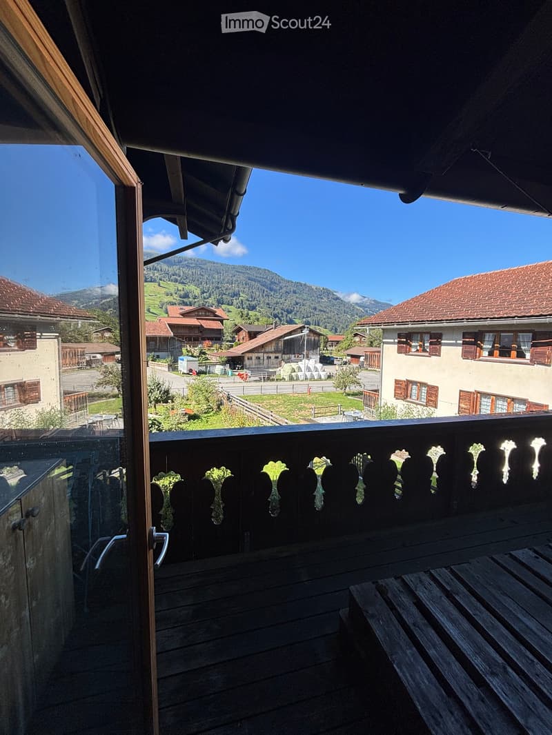 Balcony with wooden decking, ornate wooden railing, and scenic mountain view of a village with traditional houses and green fields.