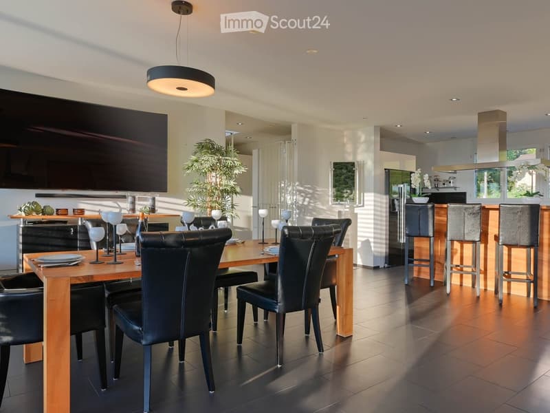Modern dining room with a wooden dining table, black leather chairs, and grey bar stools. A large flat-screen TV is mounted on the wall, and a hanging pendant light illuminates the space. The room features a sleek, contemporary design with a mix of dark a