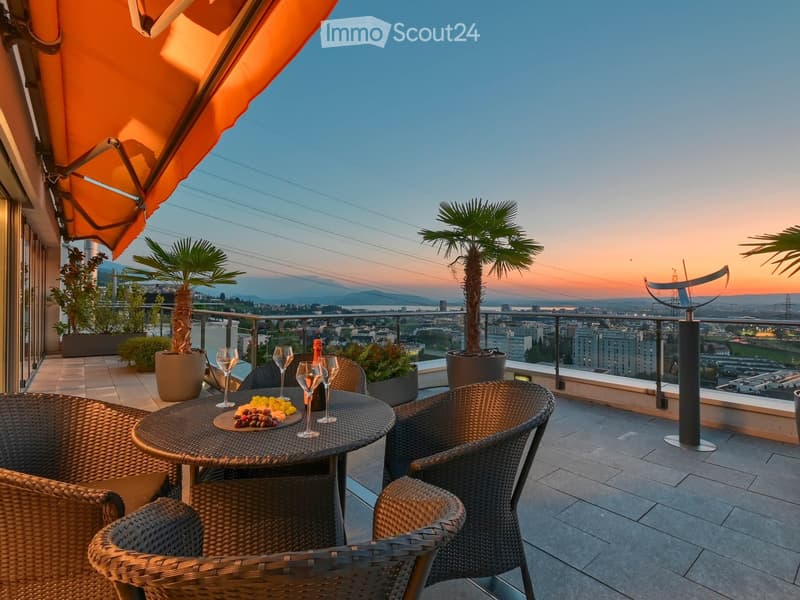 Balcony with a round table, four wicker chairs, wine glasses, a bottle, and a fruit bowl, overlooking a cityscape at sunset.