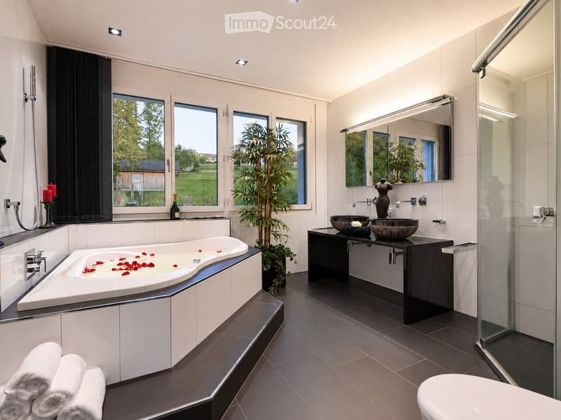 Modern bathroom with a large bathtub, double sink vanity, large mirror, and a glass-enclosed shower. Features include a potted plant, candles, and a view of greenery through large windows.