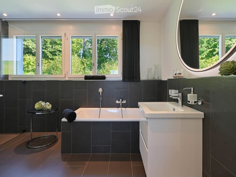 Modern bathroom with dark tiled walls, a bathtub, a sink, a large round mirror, and decorative vases on the windowsill.