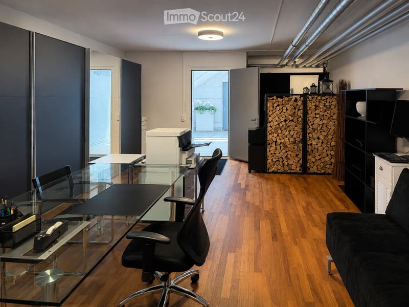 Modern office space with wooden flooring, glass desk, office chair, sofa, printer, and shelves.