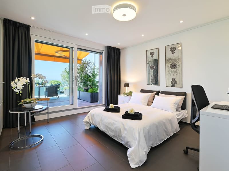 Modern bedroom with a large bed, white linens, decorative pillows, a nightstand with a lamp, a round side table with a flower arrangement, and a large window with a view of a balcony area.