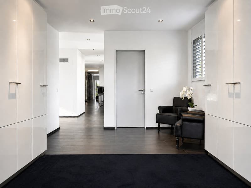 Modern hallway with white walls, dark tiled floor, and black carpeted area. Features a small seating area with a sofa, chairs, and a small table with a flower arrangement.