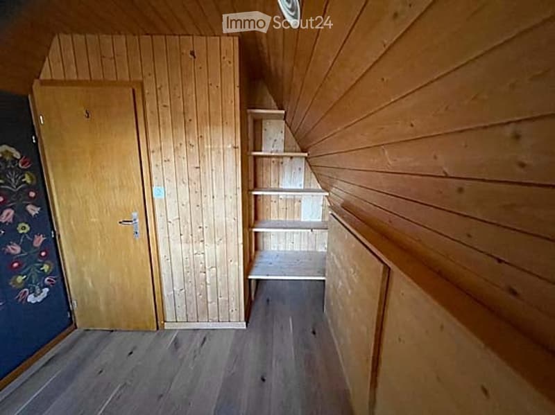 Empty wooden-walled room with a built-in shelving unit and a closed door. The space is unfurnished and features wooden flooring and walls.