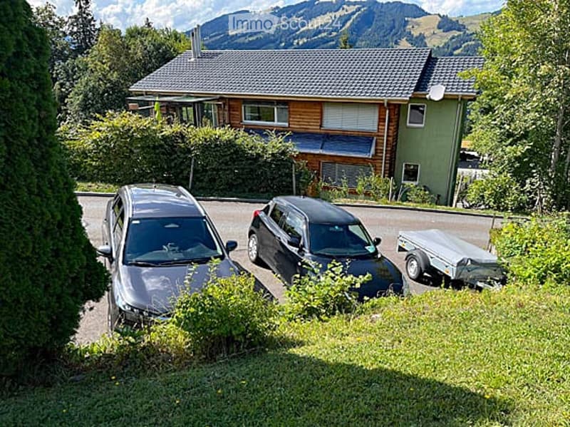 A two-story house with a tiled roof, surrounded by greenery and mountains in the background. Two cars are parked in front of the house, and there is a trailer nearby.