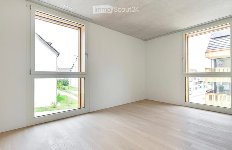 Empty room with light-colored wooden flooring, white walls, and large windows offering views of the surrounding area.