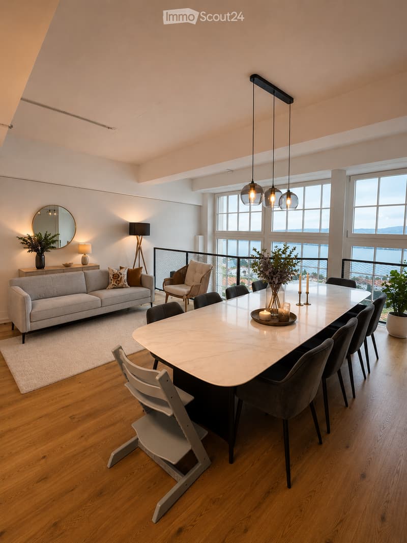 Open-concept living and dining area with a grey sofa, coffee table, dining table with chairs, pendant lights, and a view of the water through large windows and a balcony.