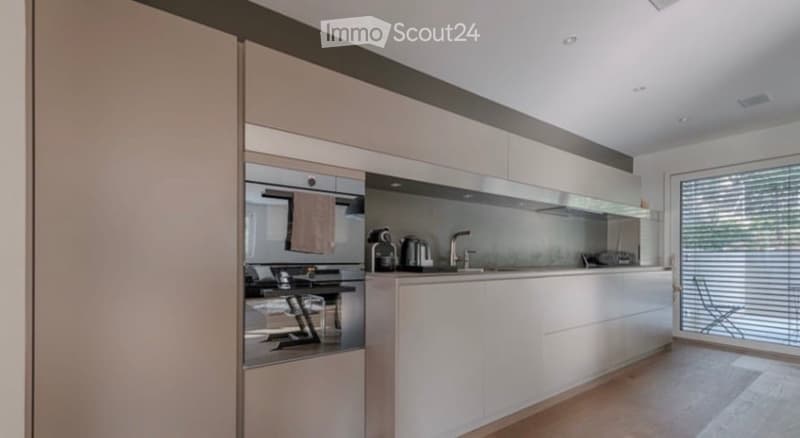 Modern kitchen with white cabinets, stainless steel appliances, and a large window with blinds. A coffee maker and oven are visible on the counter, and a chair is near the window.